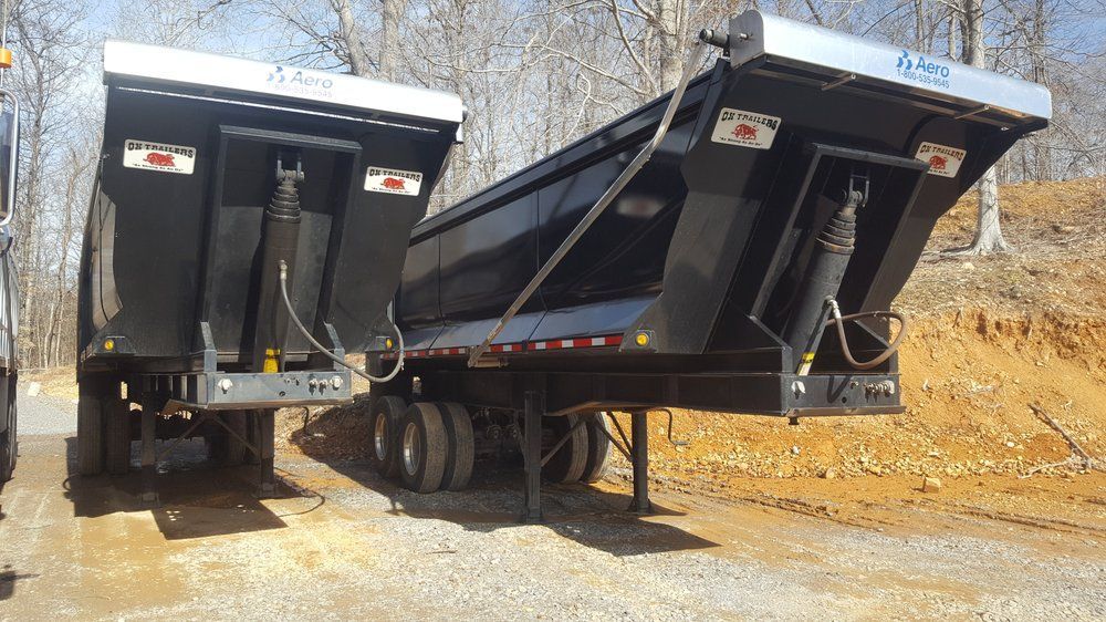Two black dump trailers parked on a gravel lot, one with its bed raised and rear gate open.