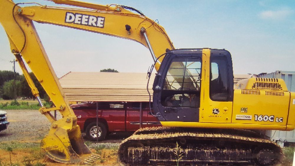 Yellow John Deere excavator parked outdoors beside a red truck and trailers under a cloudy sky