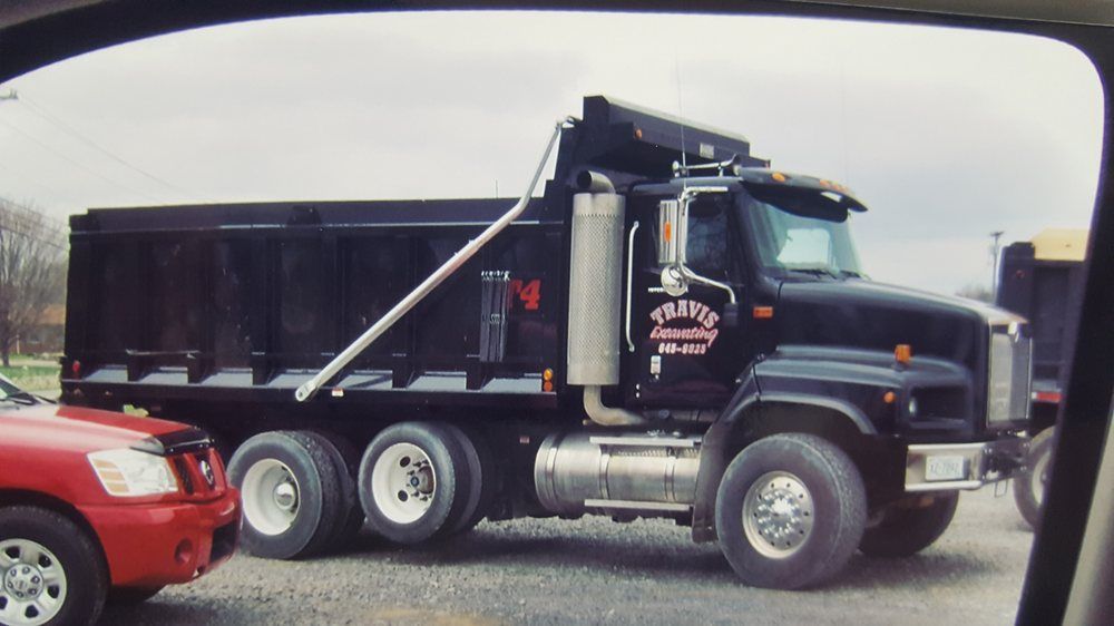 Black dump truck with raised bed parked beside a red SUV on gravel under an overcast sky