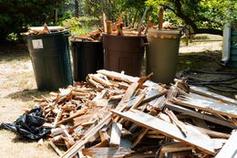 Pile of broken wooden boards beside trash bins in a yard, with a black garbage bag nearby.