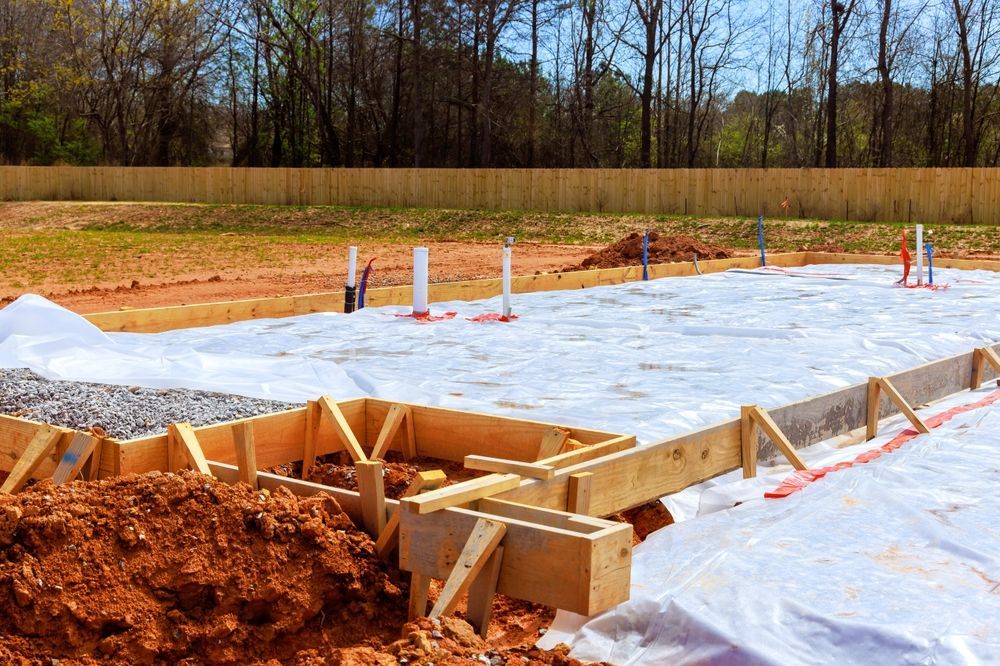 Foundation under construction on a plastic-covered site with wooden framing and orange soil piles