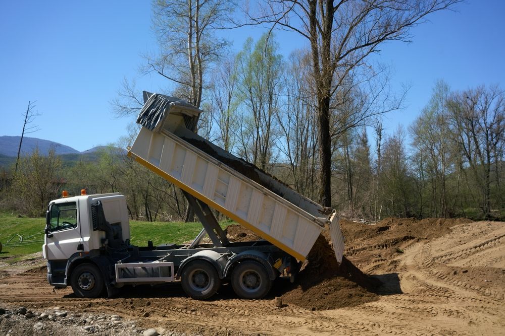 White dump truck unloads dirt at a muddy construction site with bare trees.