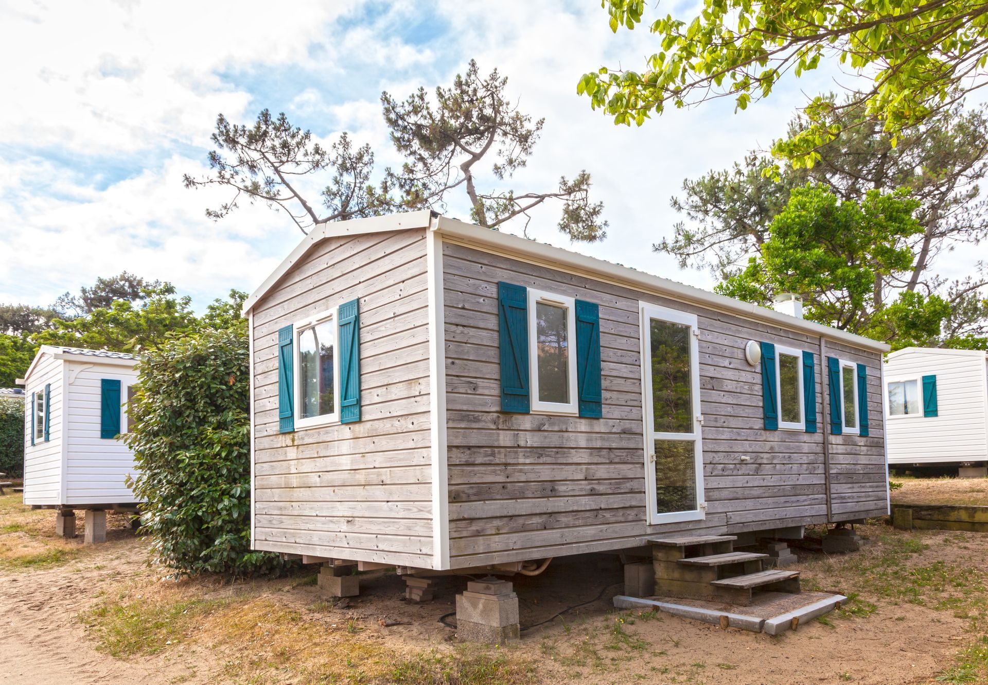 Small white cabin with blue shutters in a wooded campsite, with steps and another cabin nearby
