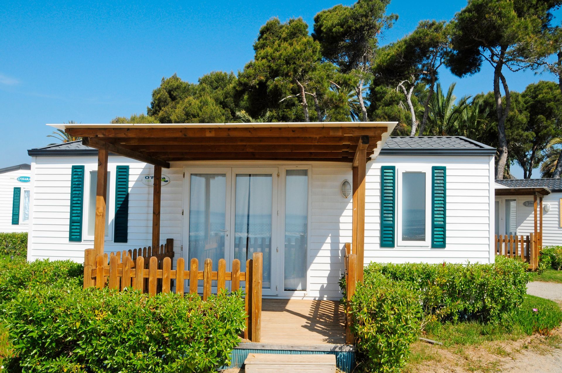 White beach cottage with teal shutters, wooden porch, and green bushes under sunny trees.