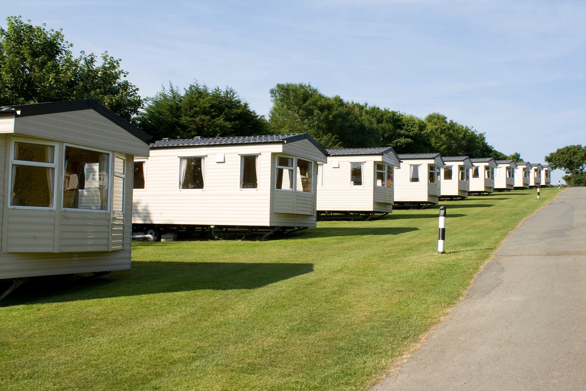 Row of cream-colored mobile homes on a grassy lot beside a paved road, under a clear sky