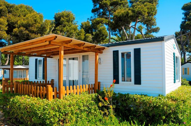 White cottage with a wooden porch and shutters, surrounded by green bushes and trees under a bright blue sky