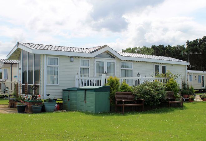 White mobile home on a grassy lot with potted plants and a green storage box under a cloudy sky