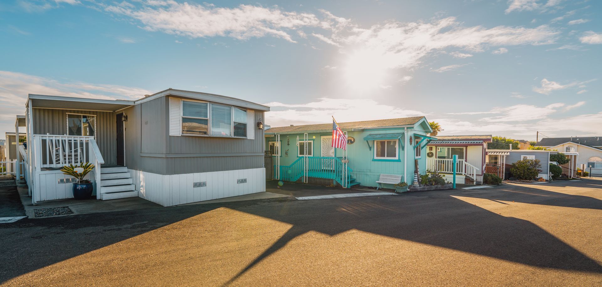Row of colorful mobile homes in a sunny trailer park with gravel lot and porch steps