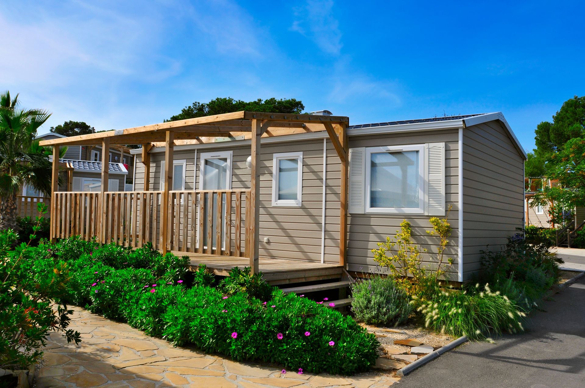 Small beige mobile home with wooden porch and garden under a blue sky
