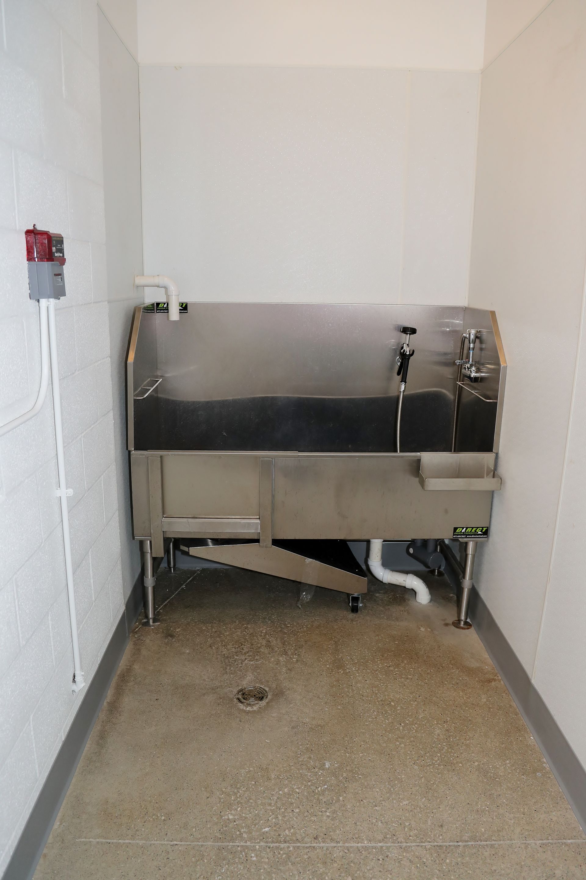 Stainless steel sink in a narrow room with concrete floor. White walls and a red emergency button.