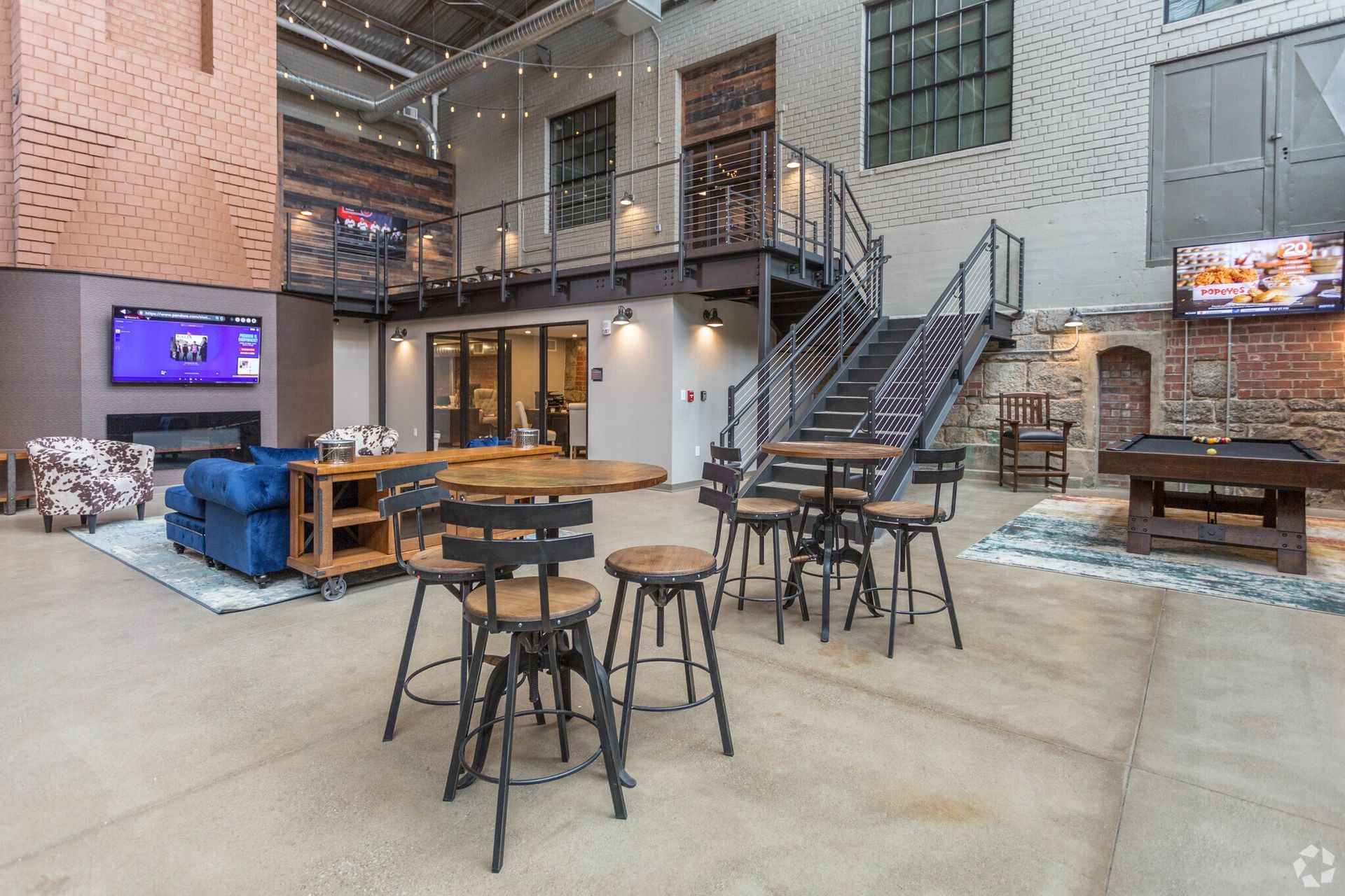 Lounge area with high-top tables, stairs to a balcony, fireplace, TV, and exposed brick walls.
