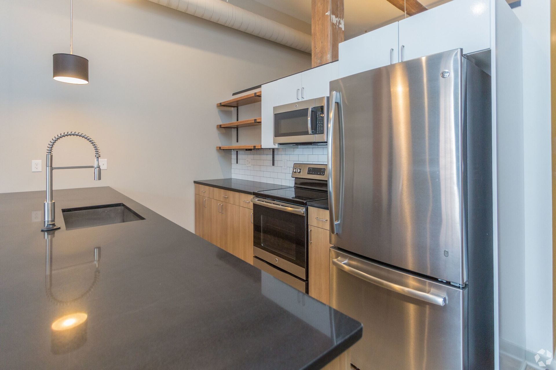 Modern kitchen with stainless steel appliances, dark countertop, light wood cabinets, and shelving.