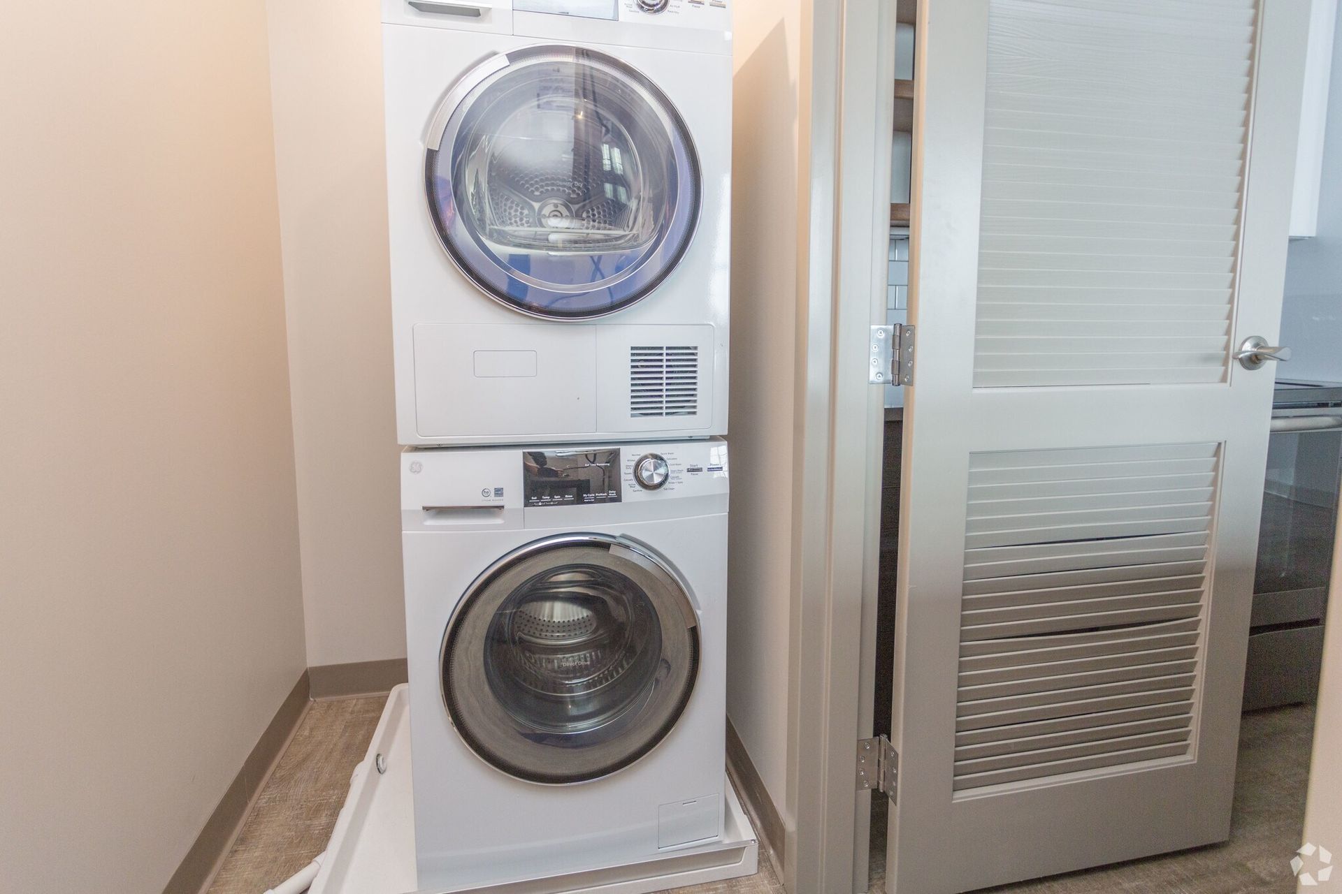 Stacked white washing machine and dryer in a utility closet with a partially open, slatted door.