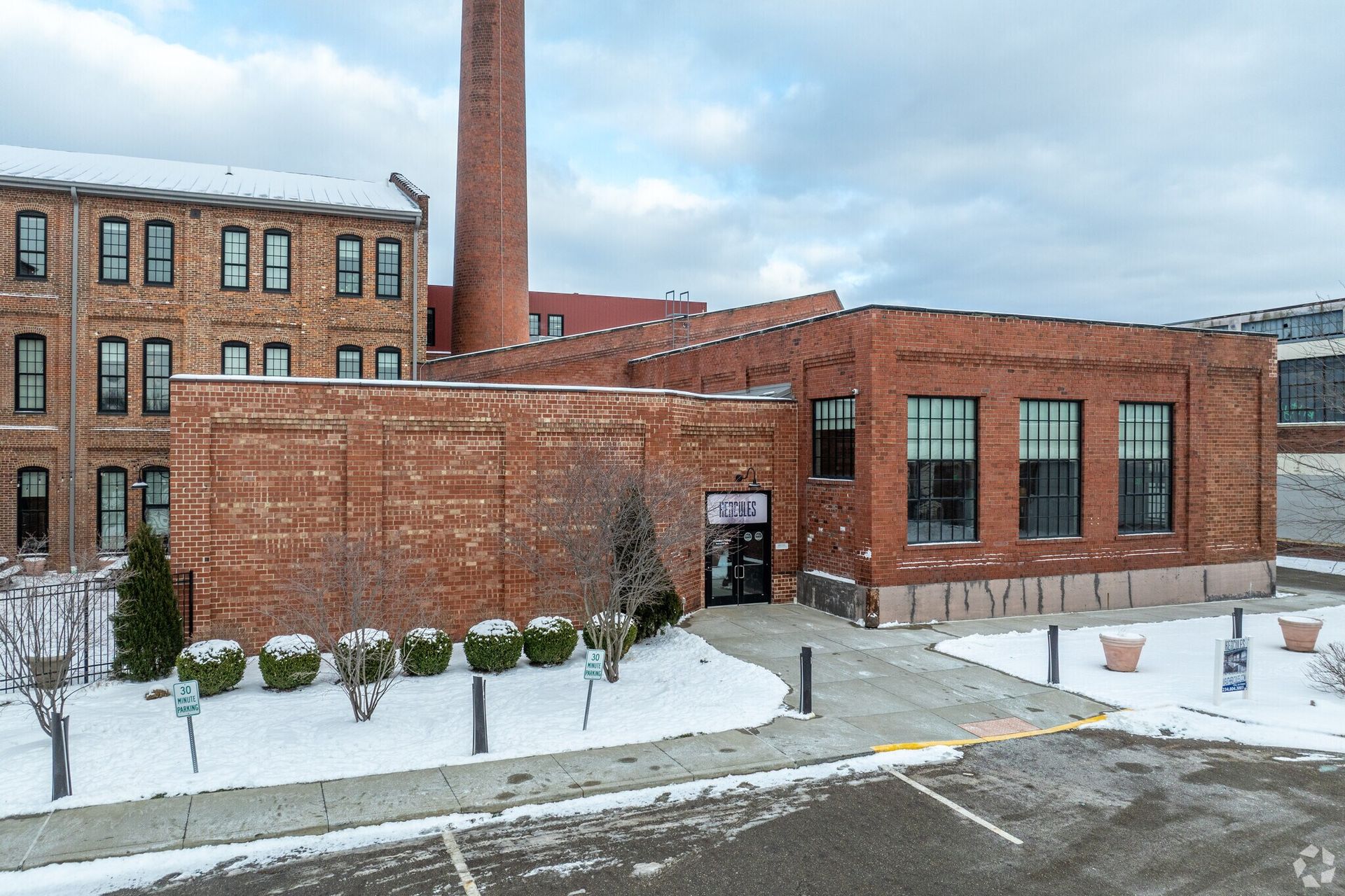 Brick industrial building with a tall smokestack, snow-covered ground, and a cloudy sky.