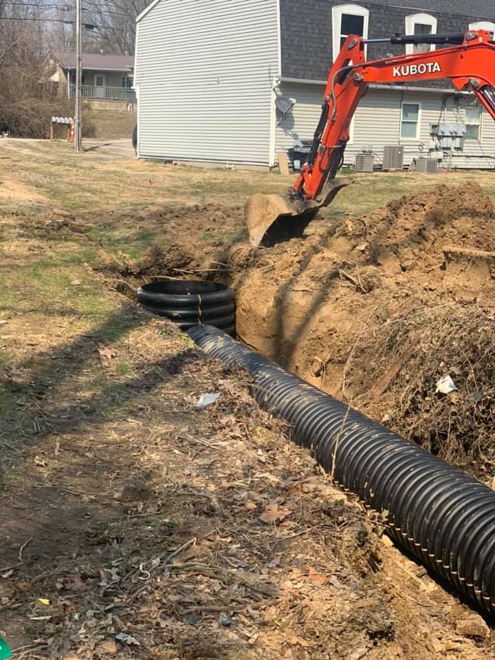 An excavator digs a trench, laying corrugated pipe near a residential home.