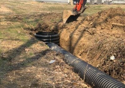 A backhoe digging a trench for a black corrugated drain pipe in a yard.