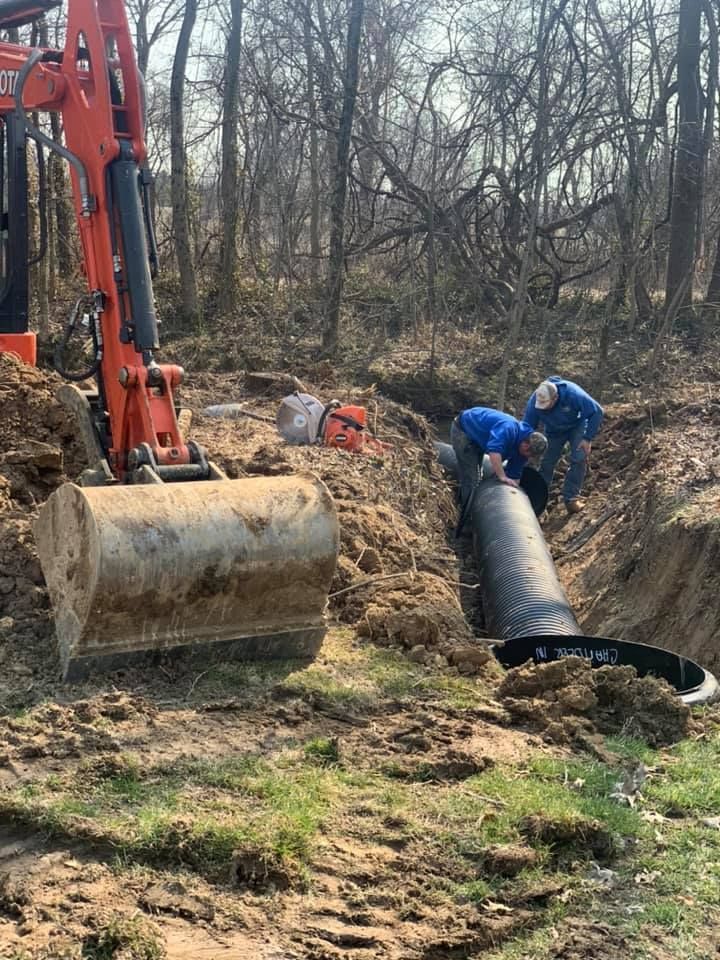 Workers installing pipe in a trench with an excavator in a wooded area.