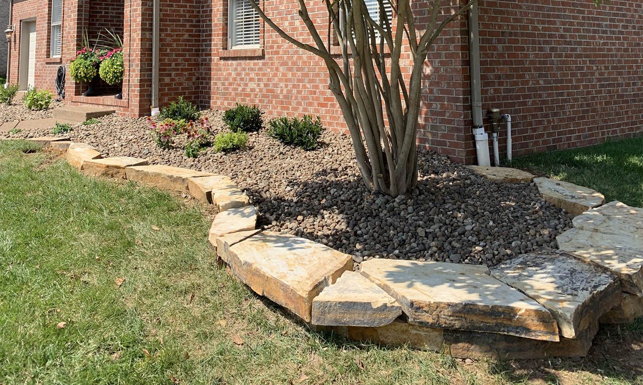 Stone-edged flower bed with small plants and brown mulch in front of a brick house, with green grass in the foreground.