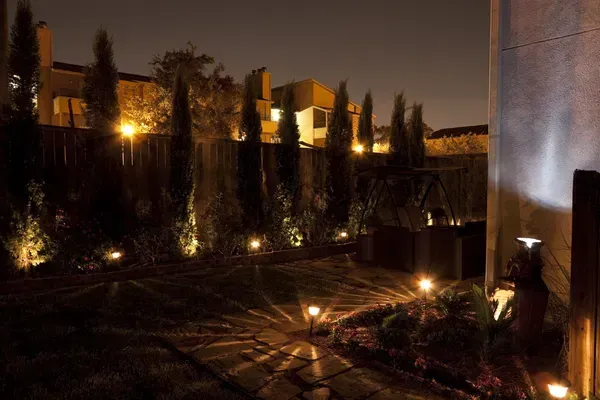 Backyard at night with spotlights illuminating trees, fence, and ground.