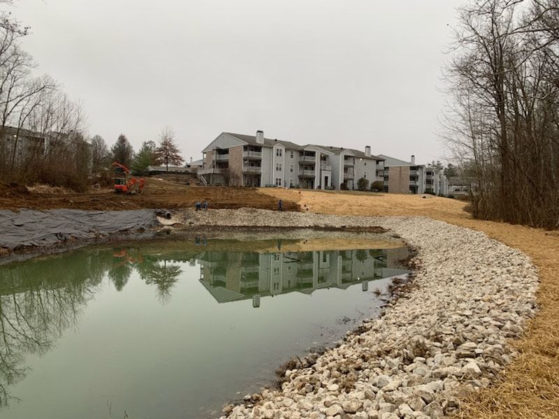 Apartment building reflected in a cloudy pond with a rock-lined edge on an overcast day.
