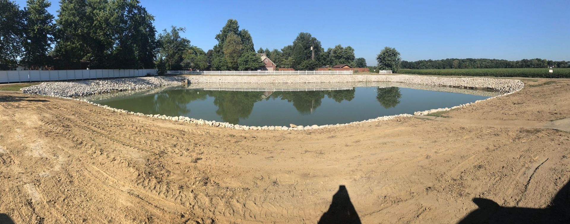 An unfinished pond with muddy banks, lined with rocks, under a clear blue sky, trees in the background.