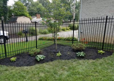 A black wrought iron fence encloses a landscaped garden bed with small plants and a tree; green grass surrounds.