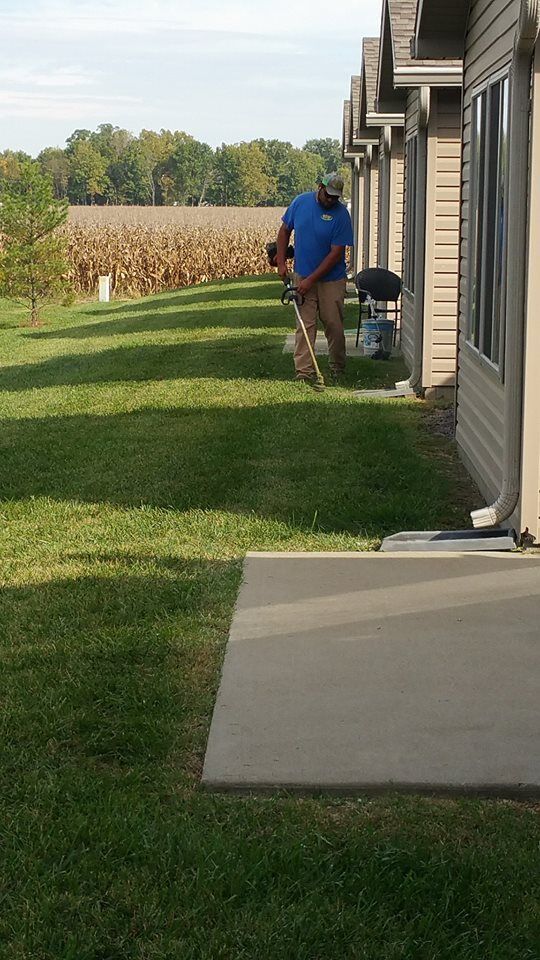 Man trimming grass with a weed wacker along a building's edge in a grassy yard on a sunny day.