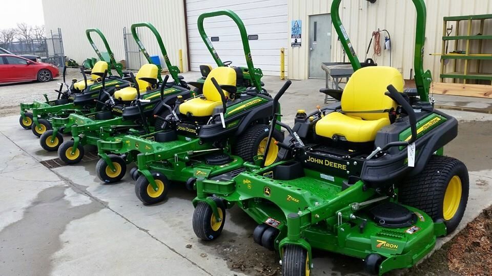 Line of green and yellow John Deere zero-turn mowers parked outside a building.