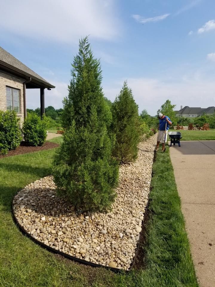 Man edging lawn beside a rock-filled garden with green trees and grass. Blue sky.
