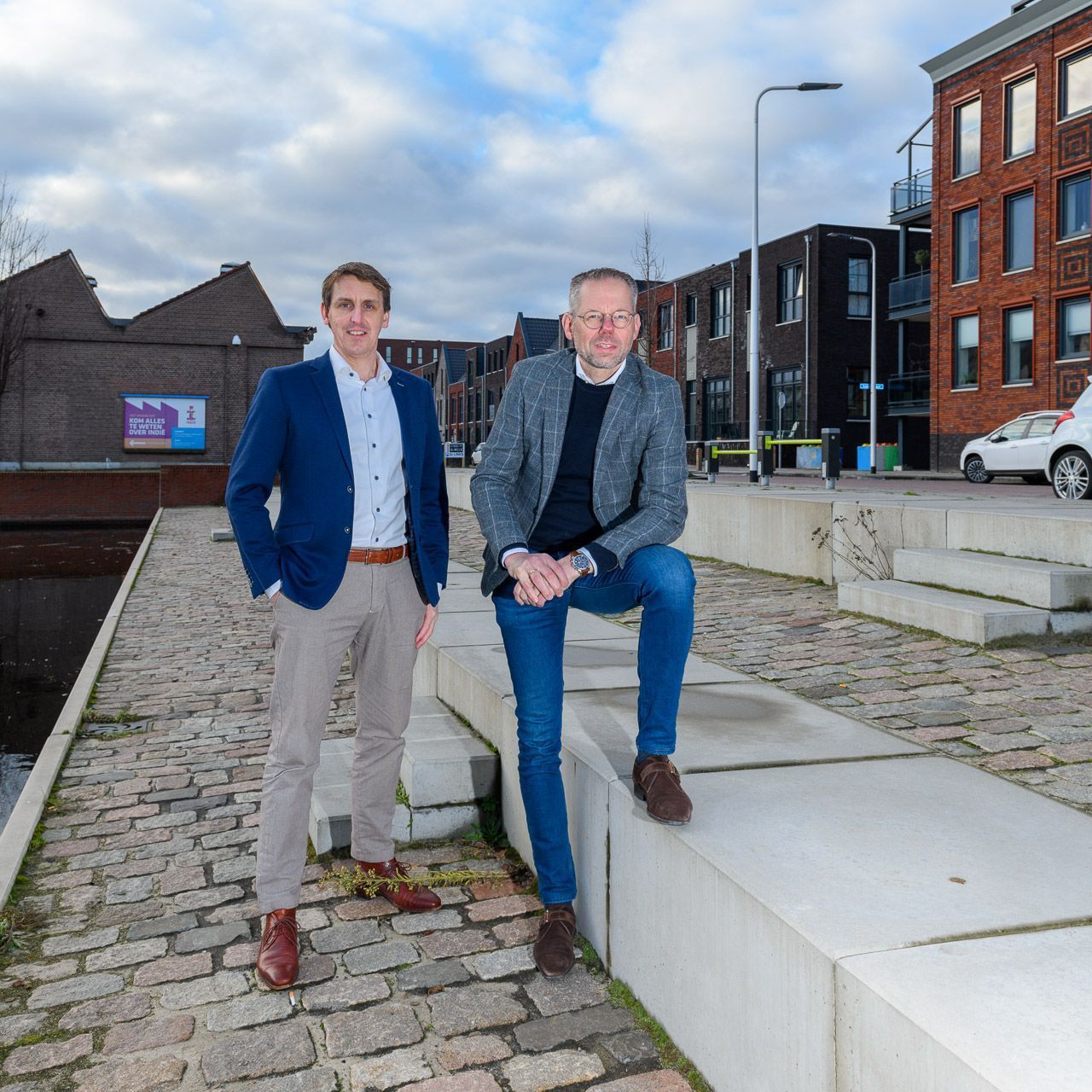 Twee mannen poseren in de buitenlucht. De ene draagt een blauwe blazer en een kaki broek, de andere een grijze jas en een spijkerbroek.