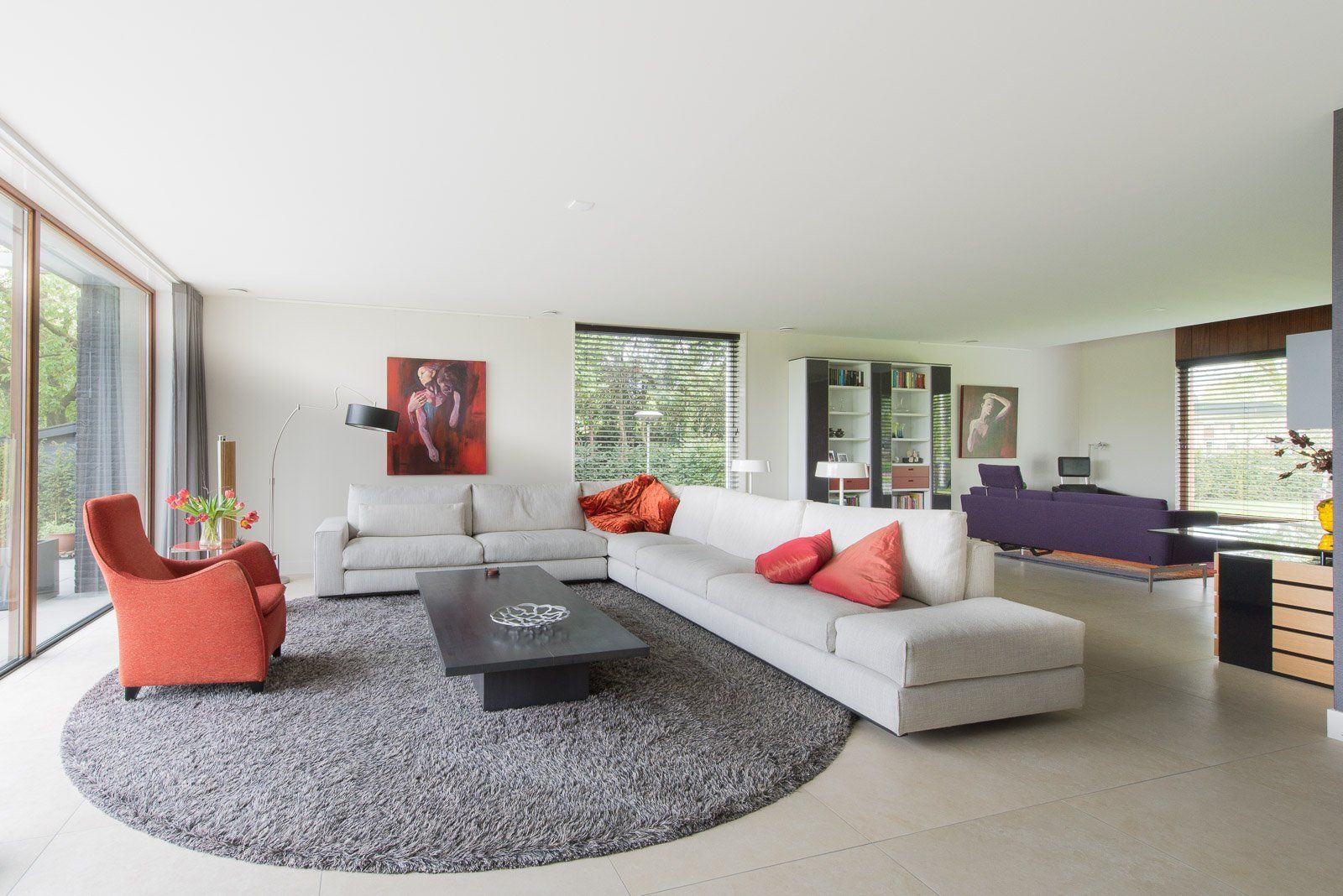 Moderne woonkamer met witte bank, grijs vloerkleed, oranje stoel en grote ramen.
