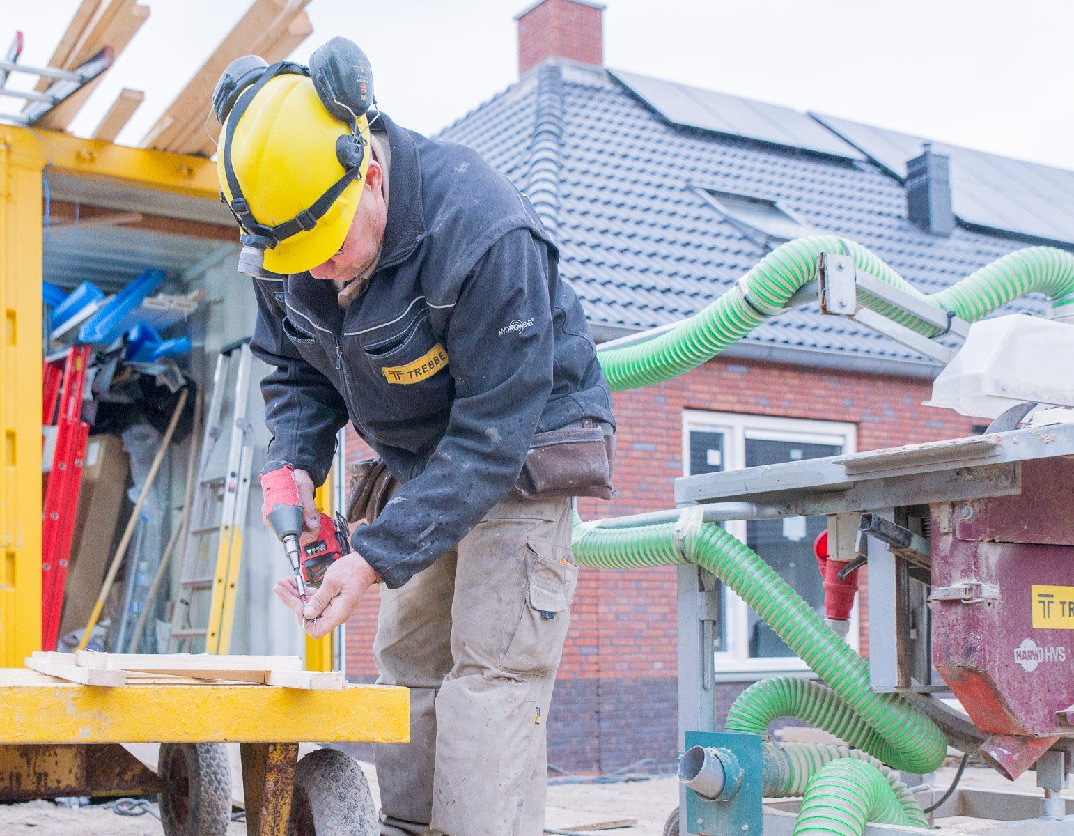 Bouwvakker met gele helm en gehoorbescherming boort in hout; buitenomgeving.