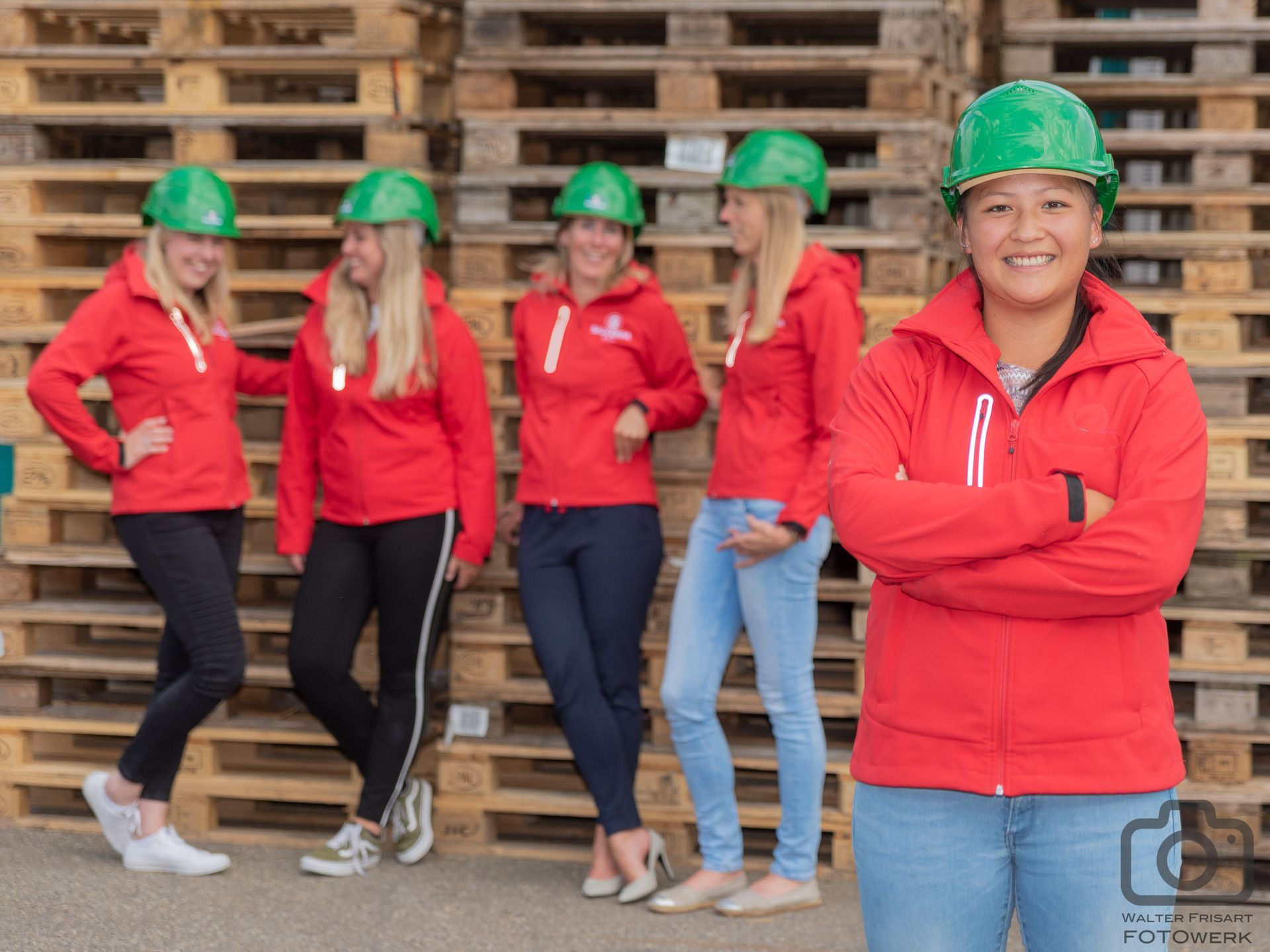 Vijf mensen in rode jassen en groene veiligheidshelmen staan voor pallets, één van hen glimlacht met de armen over elkaar.