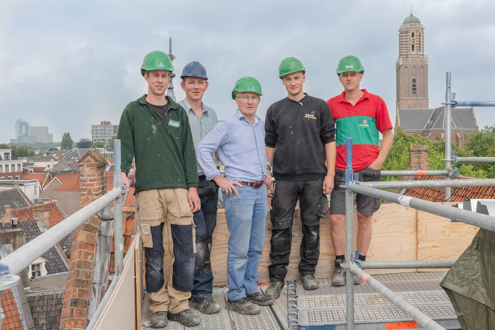 Vijf bouwvakkers met veiligheidshelmen poseren op een dak met een kerktoren op de achtergrond.