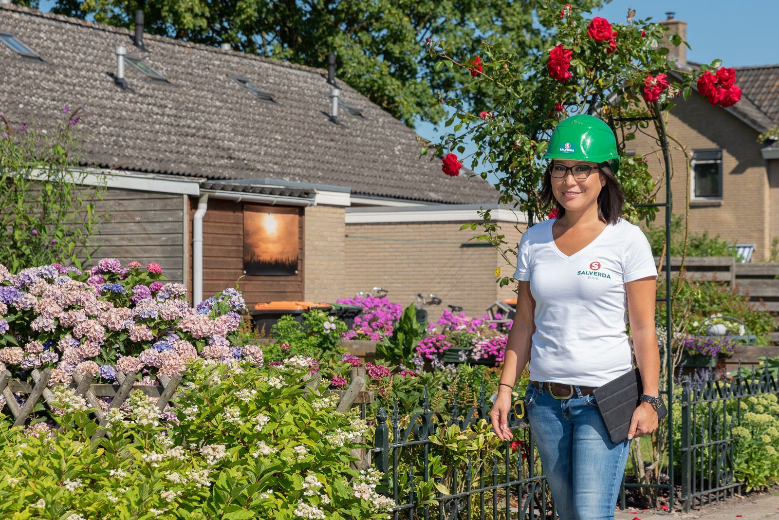 Een vrouw met een groene veiligheidshelm staat voor een huis en houdt een tablet vast; ze is omringd door een bloementuin.
