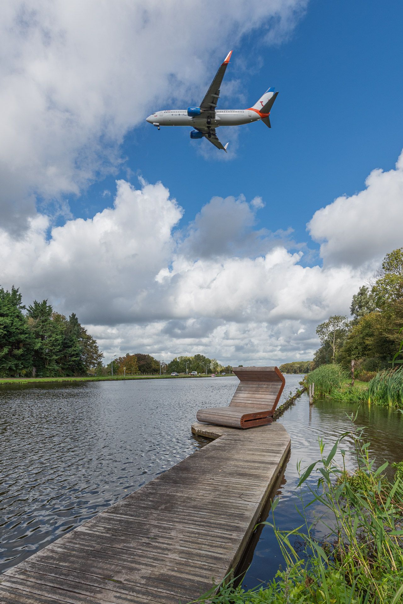 Vliegtuig vliegt boven een kanaal met een aanlegsteiger op een gedeeltelijk bewolkte dag.