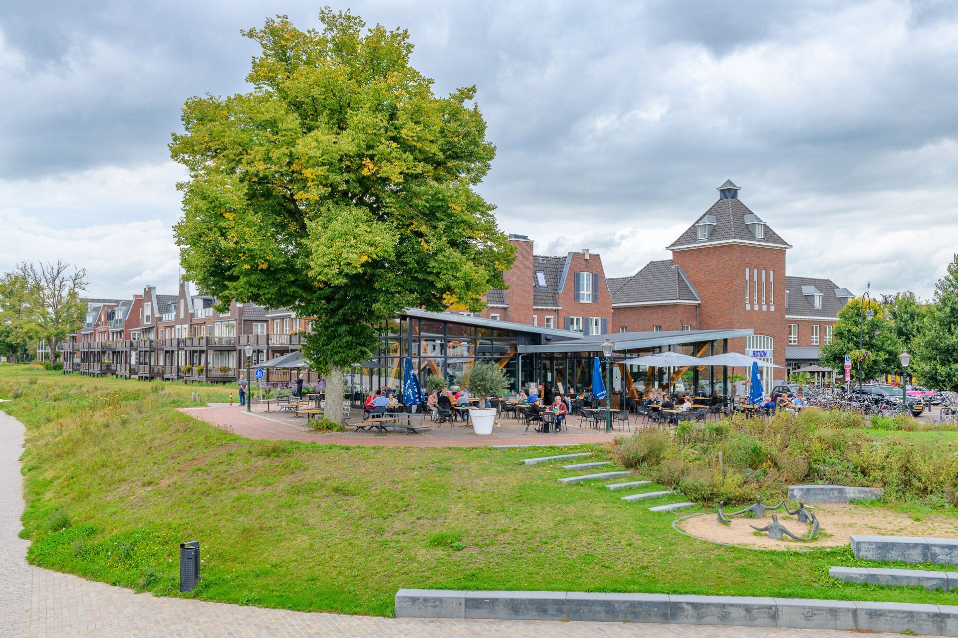 Restaurant met terras naast een grote boom en een gebouw, bewolkte dag.