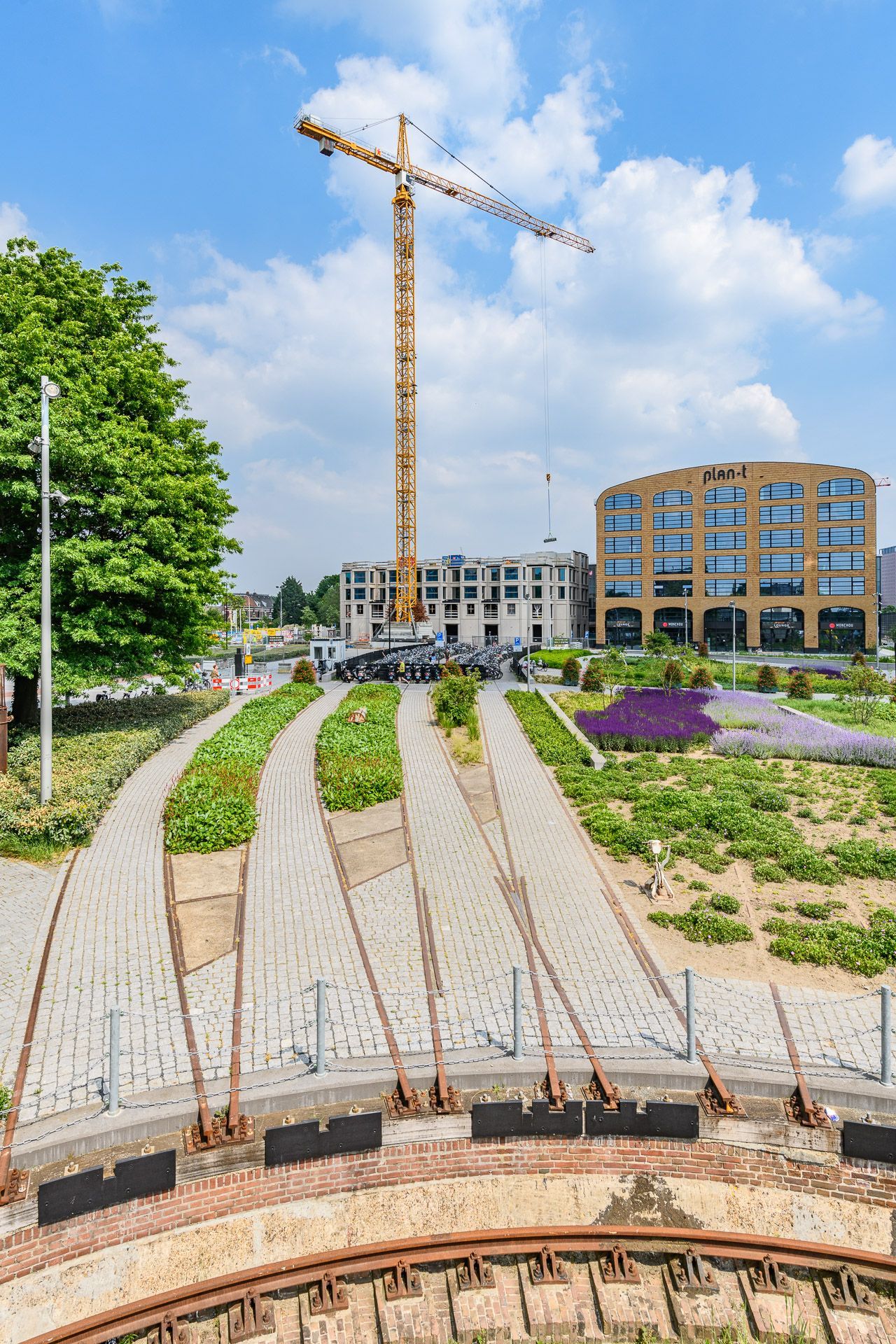 Bouwplaats met een hoge kraan; geplaveide paden leiden naar gebouwen. Zonnige dag met een blauwe lucht.