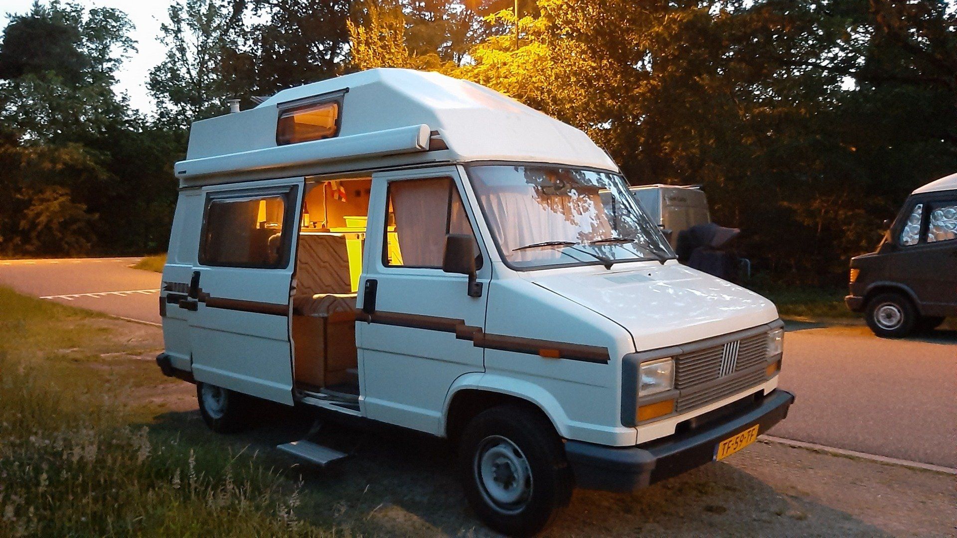 Een witte camper staat geparkeerd aan de kant van de weg, met de deur open en de binnenverlichting aan, in de schemering.