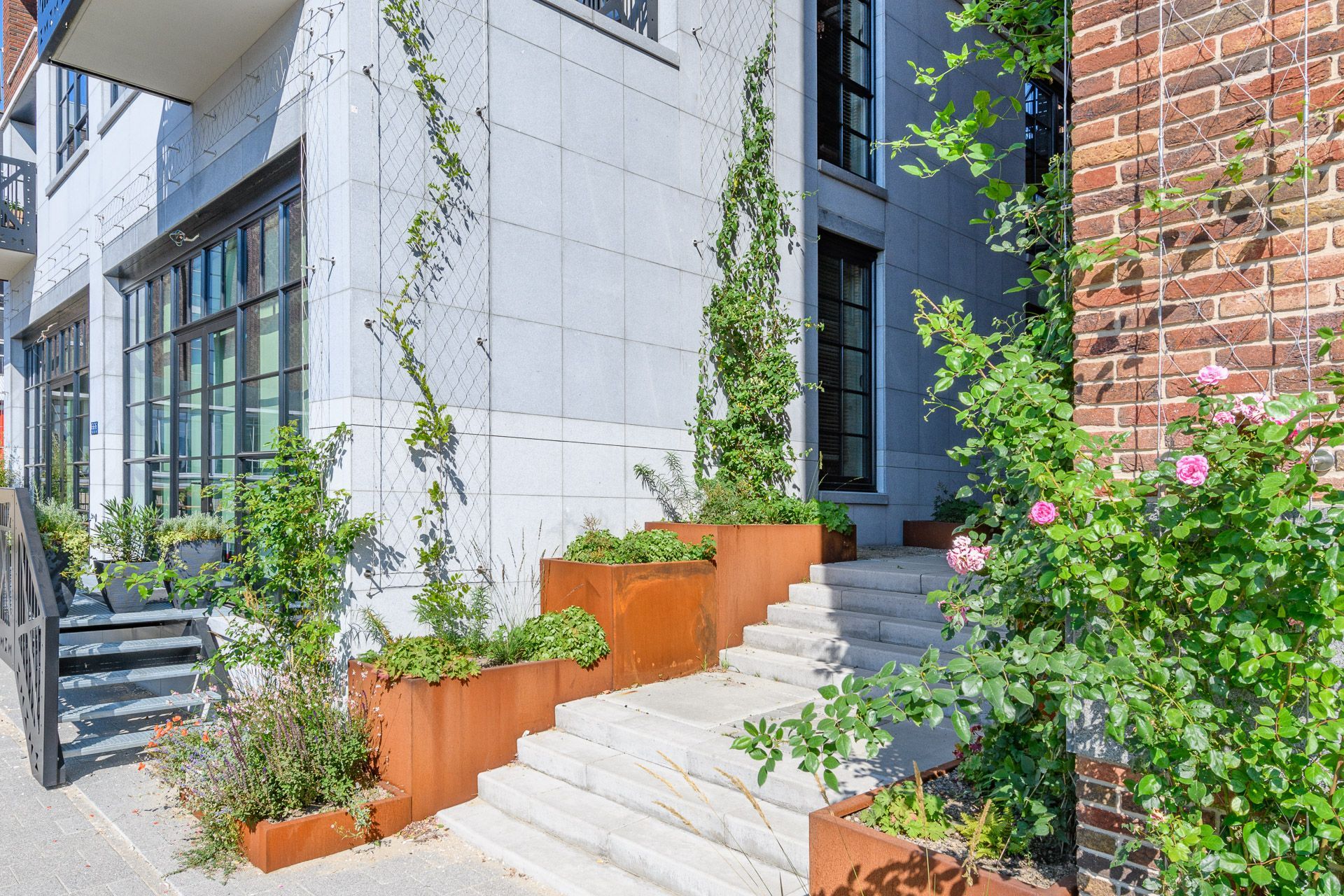 Exterieur van een modern gebouw met trappen, roestkleurige plantenbakken, groen en klimplanten.