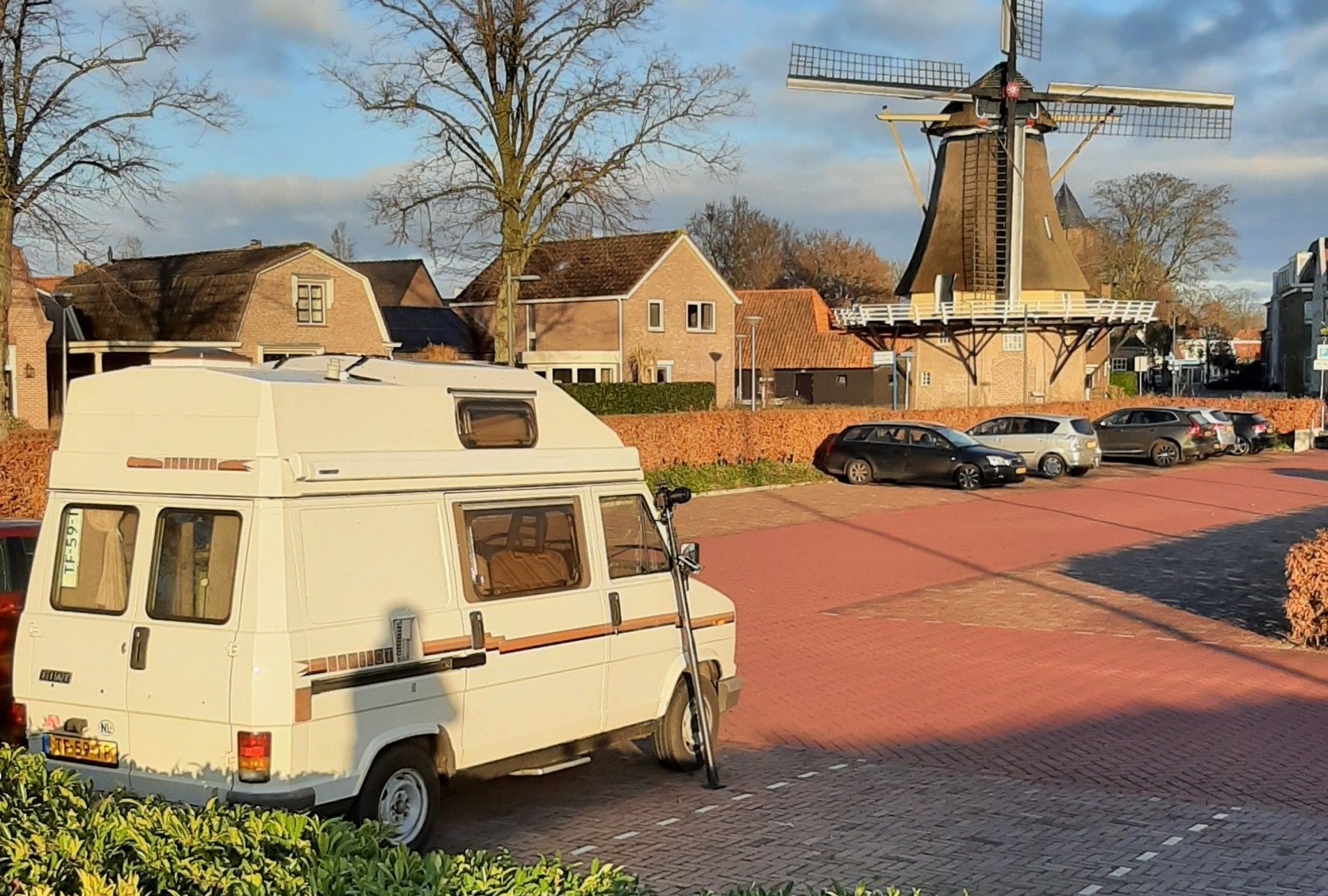 Een witte camper staat geparkeerd in een bakstenen straat, met een windmolen en huizen op de achtergrond.