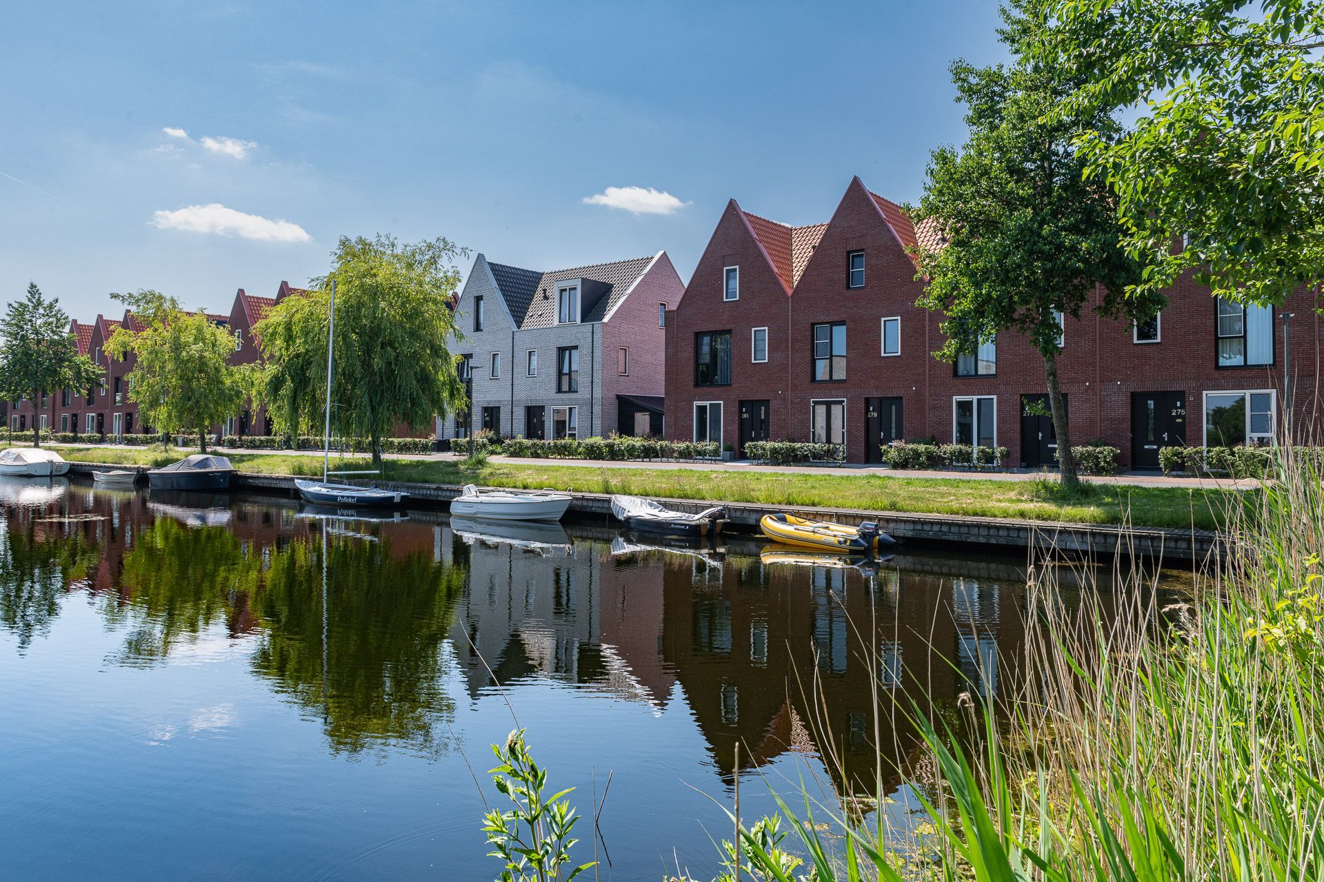 Een rij huizen langs een kanaal, met boten, water en bomen die de lucht weerspiegelen.