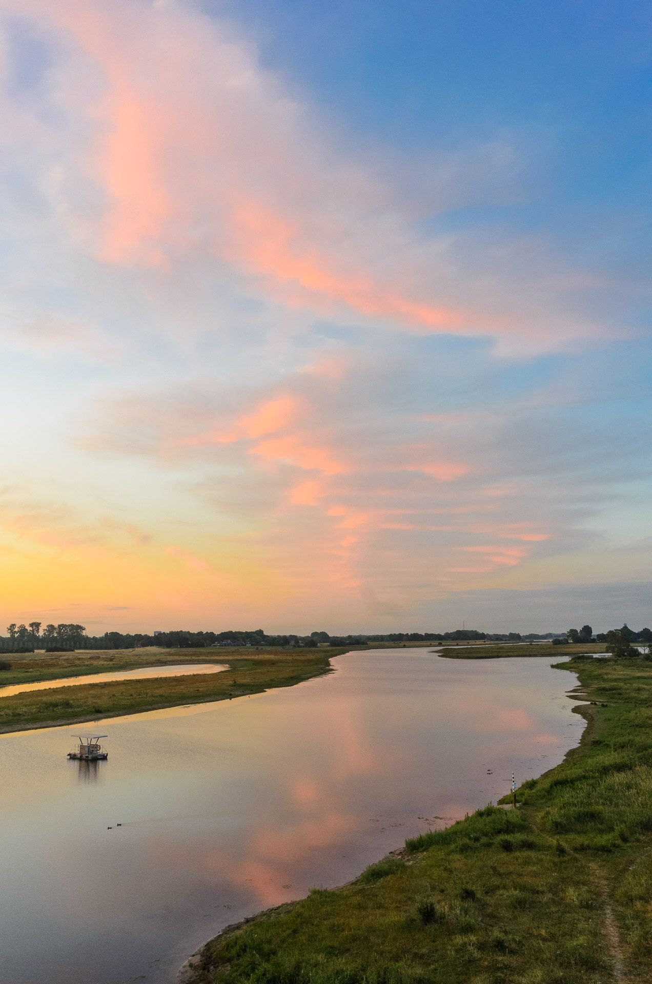 De rivier bij zonsondergang, met weerspiegeling van kleurrijke wolken. Grasrijke oevers, een klein bootje op het water.