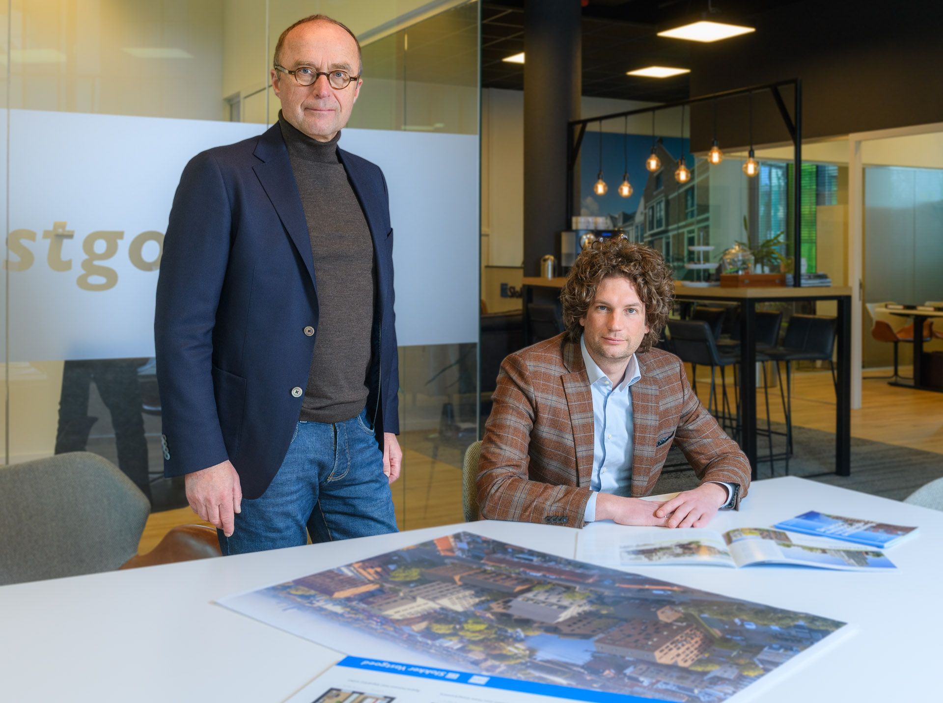 Twee mannen in een kantooromgeving; de ene staat, de andere zit aan een tafel met papieren.