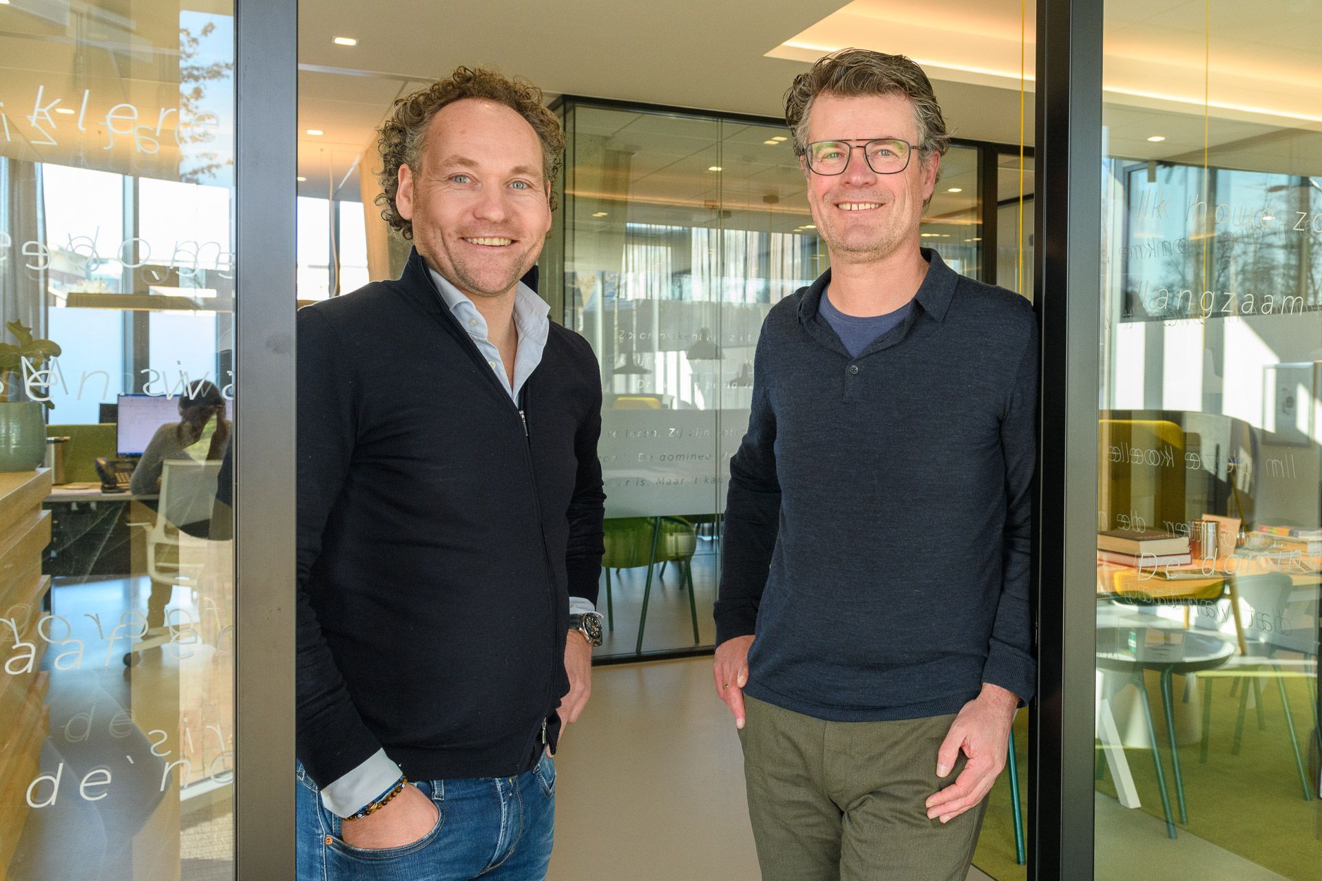 Twee mannen glimlachen in een moderne kantoordeur. De ene draagt een donkere trui, de andere een blauwe en een bril.