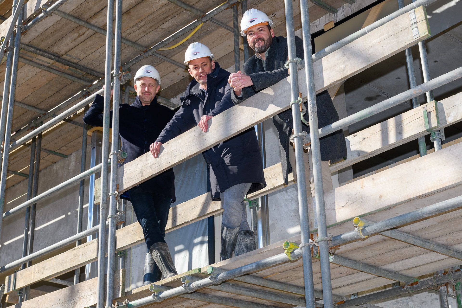 Drie mensen met veiligheidshelmen op een bouwsteiger inspecteren een gebouw.