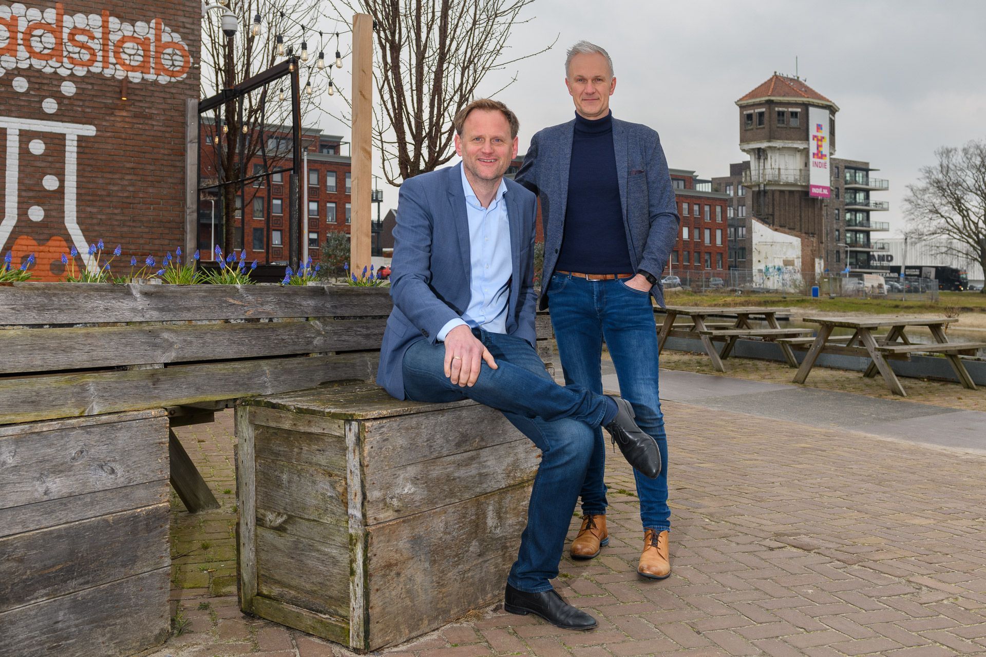Twee mannen buiten, een zit op het beton, de ander staat. Stedelijke omgeving met graffiti en een toren.