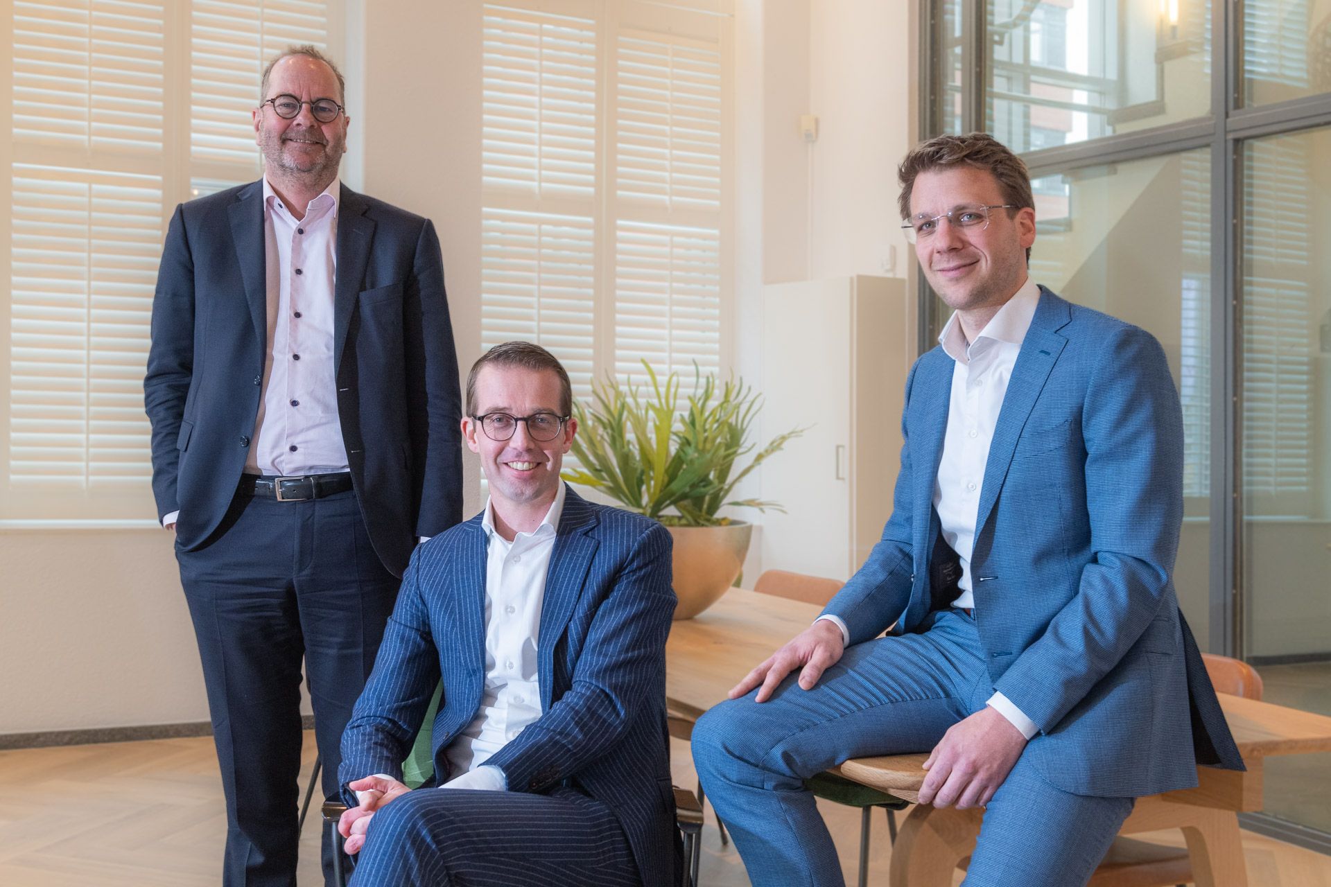 Drie mannen in pakken in een kantooromgeving. Twee zitten, één staat bij een raam.