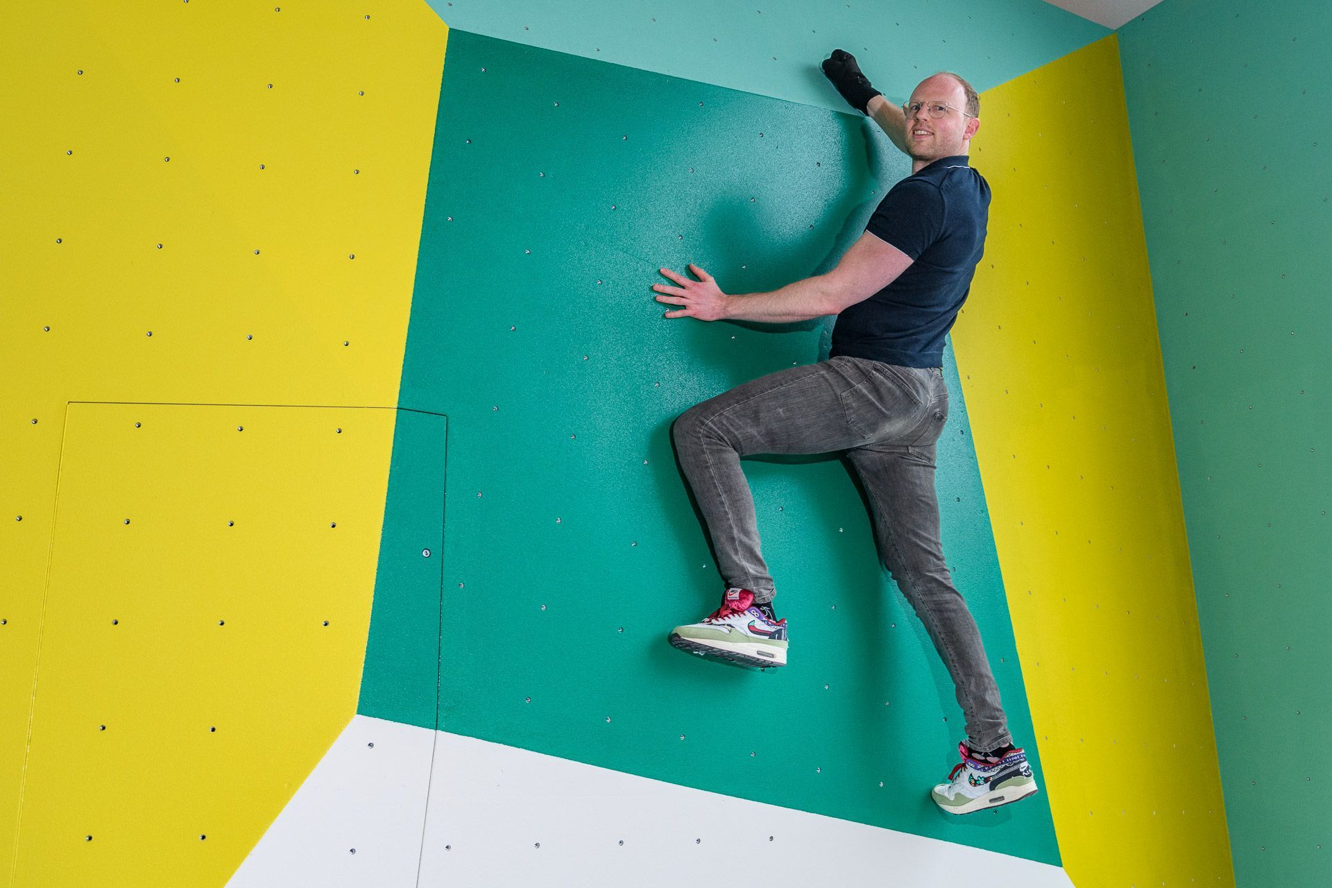 Een man beklimt een groen-gele boulderwand, gekleed in een donker shirt en een grijze broek.