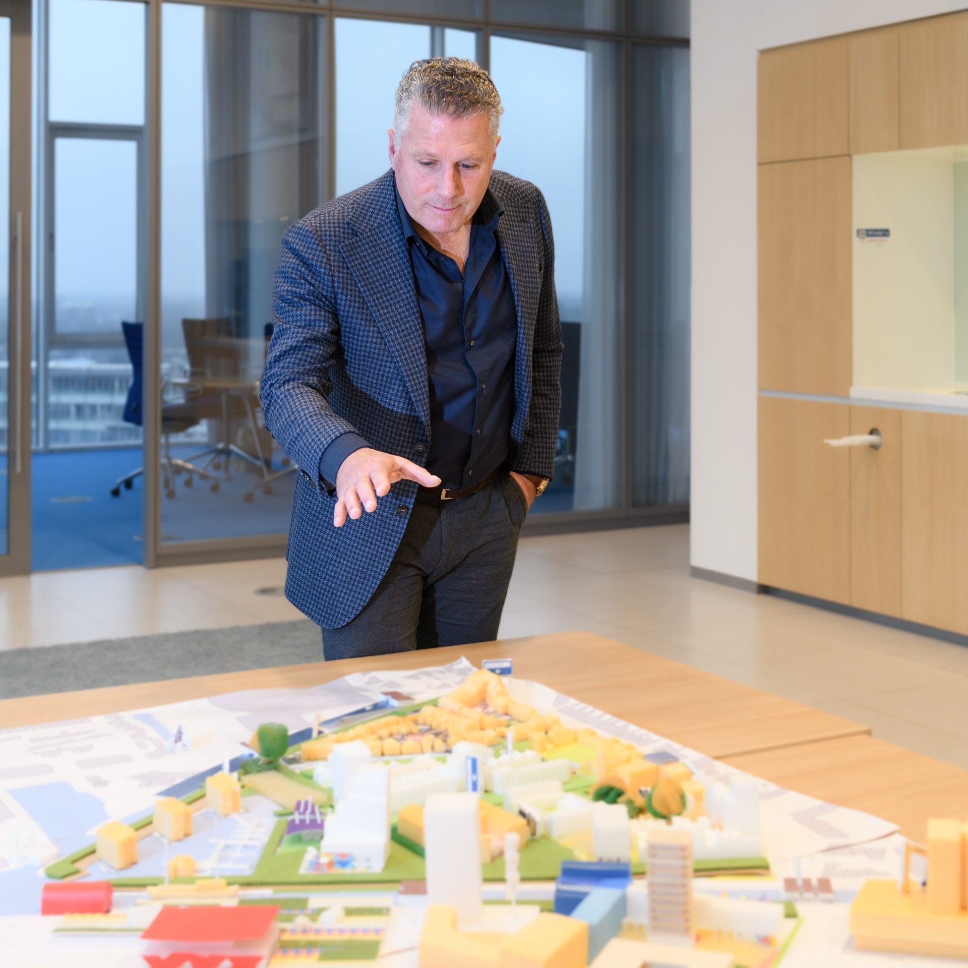 Een man in een blazer wijst naar een architectuurmodel op een tafel in een kantooromgeving.